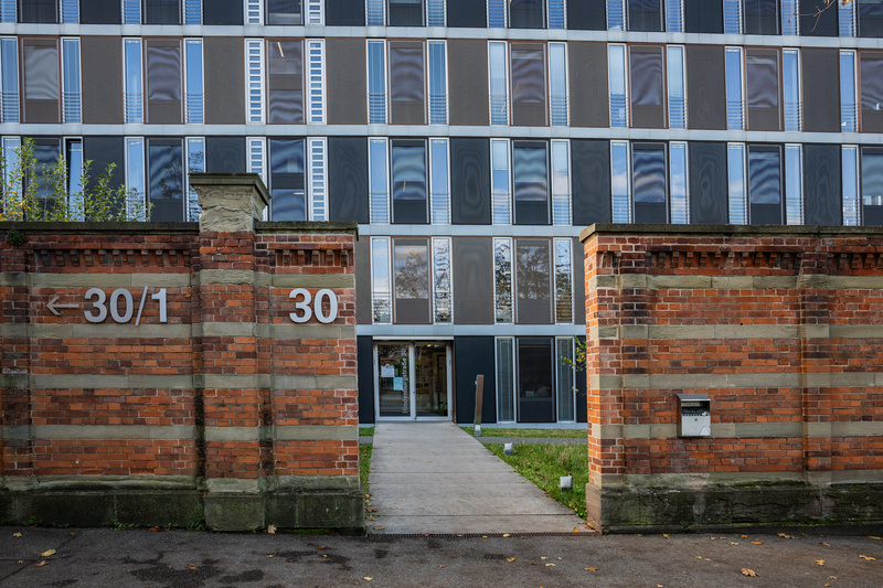 Ein moderner Eingang zu einem Gebäude mit großen Fenstern. Die Fassade besteht aus einer Kombination von grauen und blauen Paneelen. Rechts und links vom Eingang sind massive, rote Backsteinmauern mit großen Ziffern "30" und "30/1" angebracht.