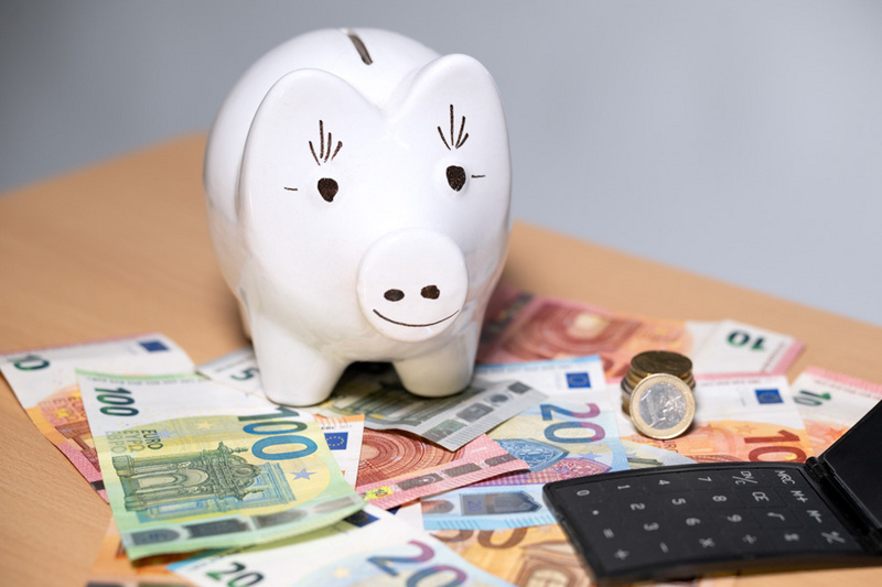 Ein Sparschwein aus Keramik steht auf einem Tisch, umgeben von verschiedenen Geldscheinen in Euro, darunter 20- und 100-Euro-Scheine. Neben dem Sparschwein liegt eine Münze und eine schwarze Geldbörse. Die Szene vermittelt das Thema Sparen und Geldverwaltung.