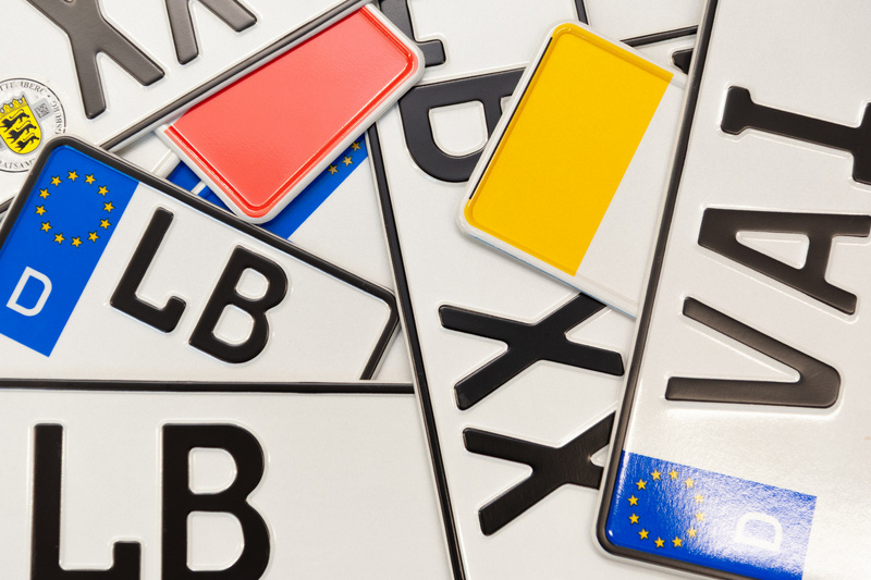 Mehrere Autokennzeichen liegen übereinander, darunter sind die Buchstabenkombinationen "LB" und "VAI" sowie europäische Abzeichen zu sehen. Einige der Schilder haben unterschiedliche farbige Rahmungen in Rot und Gelb. (Text KI generiert)