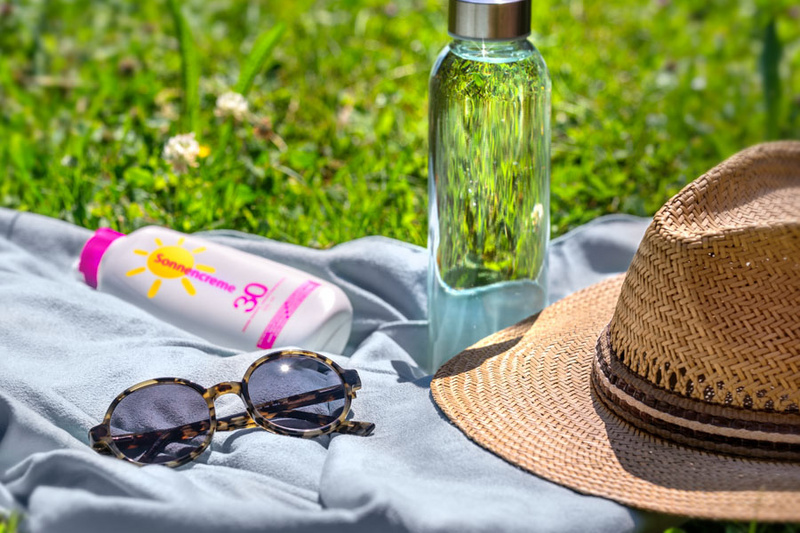 Ein Handtuch liegt auf grünem Gras, darauf befinden sich eine Flasche Wasser, eine Sonnencreme mit gelbem Etikett und eine Sonnenbrille. Neben diesen Gegenständen liegt ein Strohhut. Die Szene vermittelt ein entspanntes Sommergefühl.