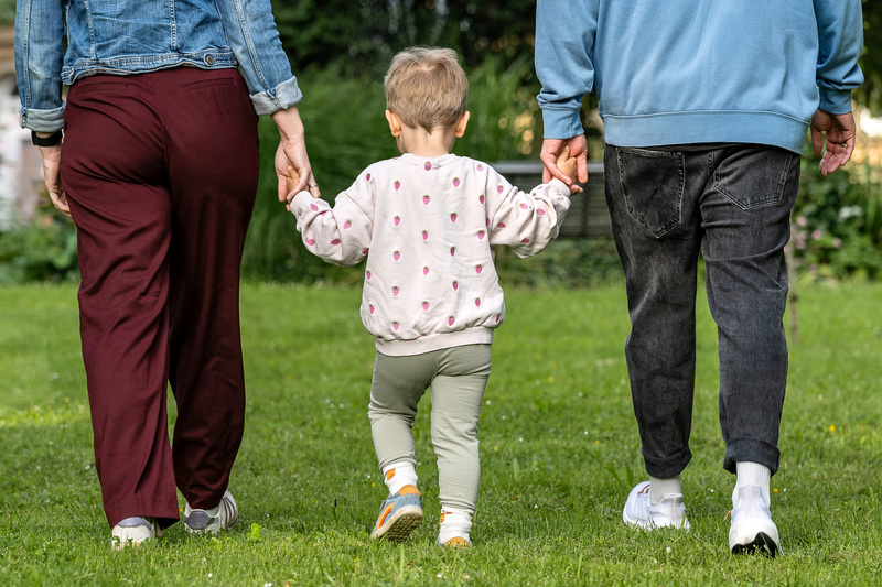 Ein Kind hält die Hände von zwei Erwachsenen und geht auf einer grünen Wiese. Die Person auf der linken Seite trägt eine Jeansjacke und rote Hose, die Person auf der rechten Seite trägt einen blauen Pullover und schwarze Jeans. Das Kind trägt ein gepunktetes Sweatshirt und grüne Hosen. (Text KI generiert)