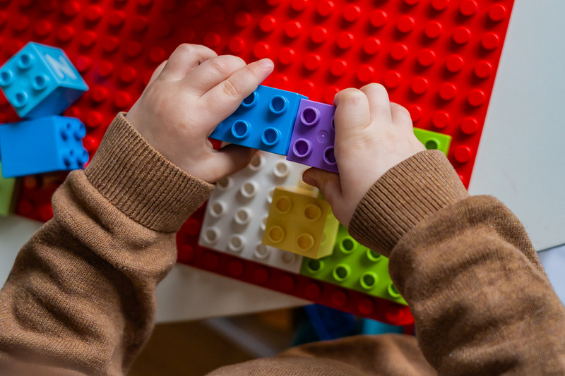 Ein Kind baut mit bunten Legosteinen auf einer roten Basisplatte. Die Hände des Kindes halten zwei miteinander verbundene Steine, während weitere Steine in verschiedenen Farben um sie herum liegen. (Text KI generiert)