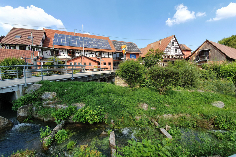 Eine malerische Ansicht eines Gebäudekomplexes mit Solarpanelen auf dem Dach, umgeben von grüner Vegetation und einem kleinen Fluss. Eine Brücke führt über den Fluss, während im Hintergrund weitere traditionelle Gebäude zu sehen sind. Der Himmel ist blau mit einigen Wolken. (Text KI generiert)
