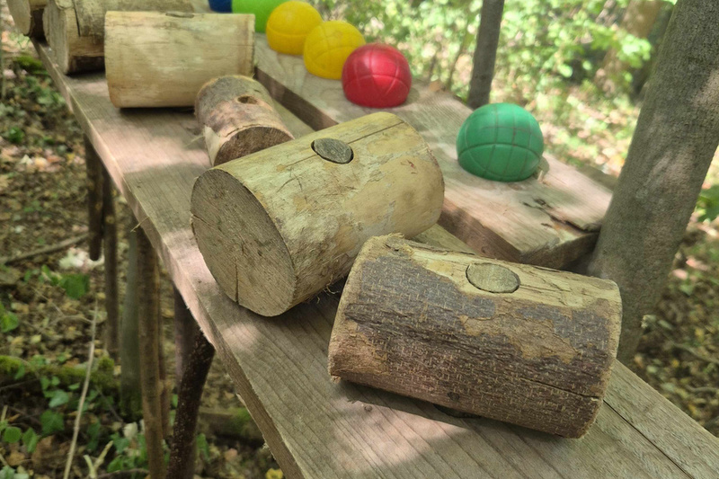 Auf einer Holztischplatte im Wald liegen mehrere Baumstämme und bunte Kugeln in den Farben Blau, Gelb, Rot und Grün. Der Tisch ist von Bäumen umgeben und der Boden ist mit Laub bedeckt. (Text KI generiert)