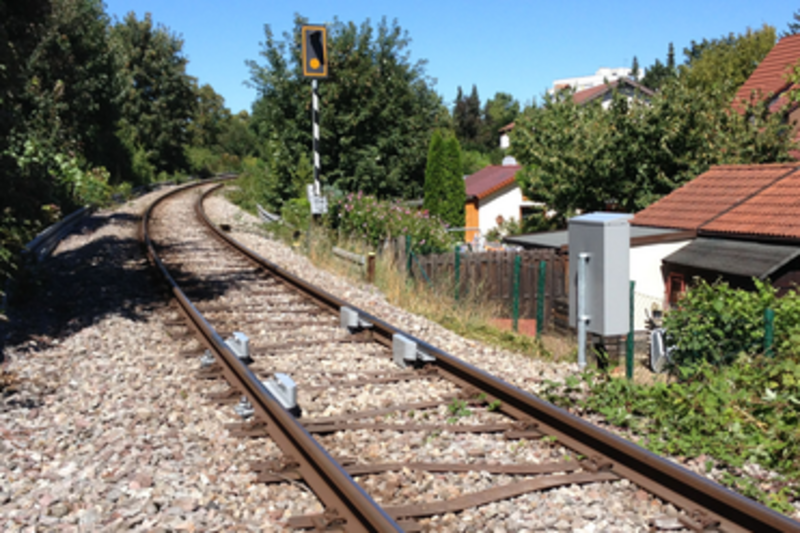Blick auf eine kurvige Bahnstrecke, die von Schienen und Kies umgeben ist. Im Hintergrund sind Bäume und Wohnhäuser sichtbar. Ein Signal steht am Rand der Gleise. (Text KI generiert)