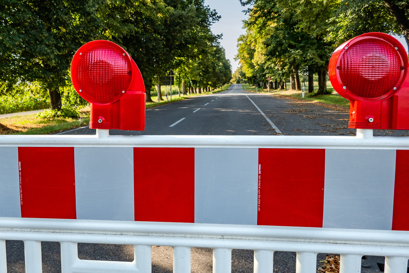 Eine Straßenbarriere mit roten Blinklichtern und weißen sowie roten Abschnitten ist im Vordergrund zu sehen. Im Hintergrund erstreckt sich eine leere Straße, die von Bäumen gesäumt ist, die im Sonnenlicht stehen. Die Barriere blockiert den Zugang zur Straße.
