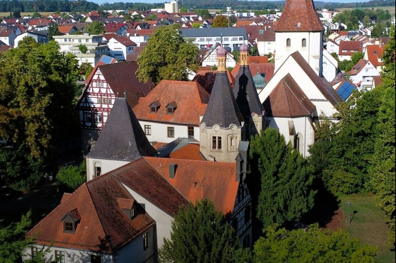 Entdecke unsere Region - 1 Luftaufnahme eines historischen Gebäudes in Hemmingen, umgeben von Bäumen und Wohnhäusern. Im Vordergrund sind ein Rathaus und ein Kirchturm sichtbar. Darüber steht der Text „Hemmingen ENTDECKE UNSERE REGION“. (Text KI generiert)