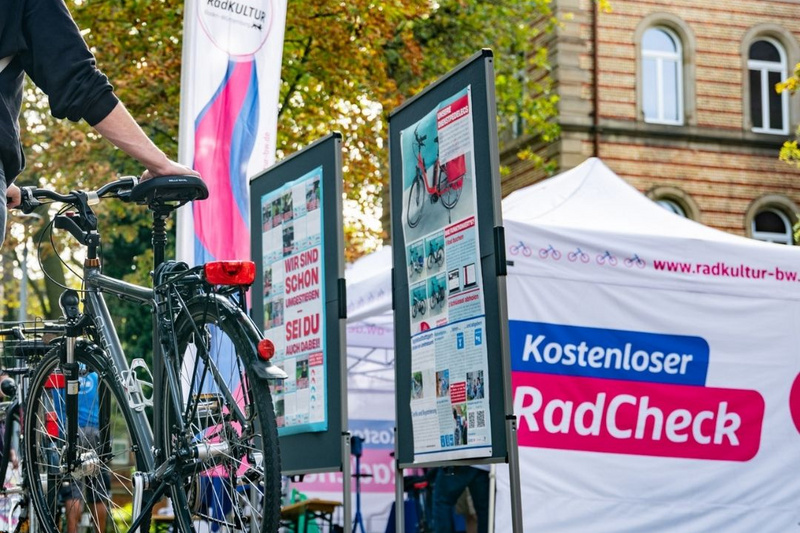Veranstaltungen: Facebook, Story, Website - 5 Eine Person steht neben einem Fahrrad auf einem herbstlichen Platz, umgeben von buntem Laub. Im Hintergrund sind Informationsstände mit Plakatwänden und ein Zelt, das für einen kostenlosen Radcheck wirbt. Die Atmosphäre ist freundlich und einladend. (Text KI generiert)
